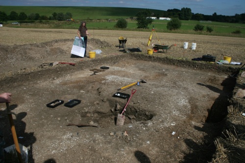 Trench with well-like feature in the foreground (quartered for excavation) and the dark grey line of the flanking ditch of the trackway at the far end of the trench